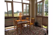A small sunroom or enclosed porch with large windows showing a green outdoor view. Inside, there is a wooden table with two chairs, a table lamp, and a decorative rug on the floor. The room has a cozy and inviting atmosphere.