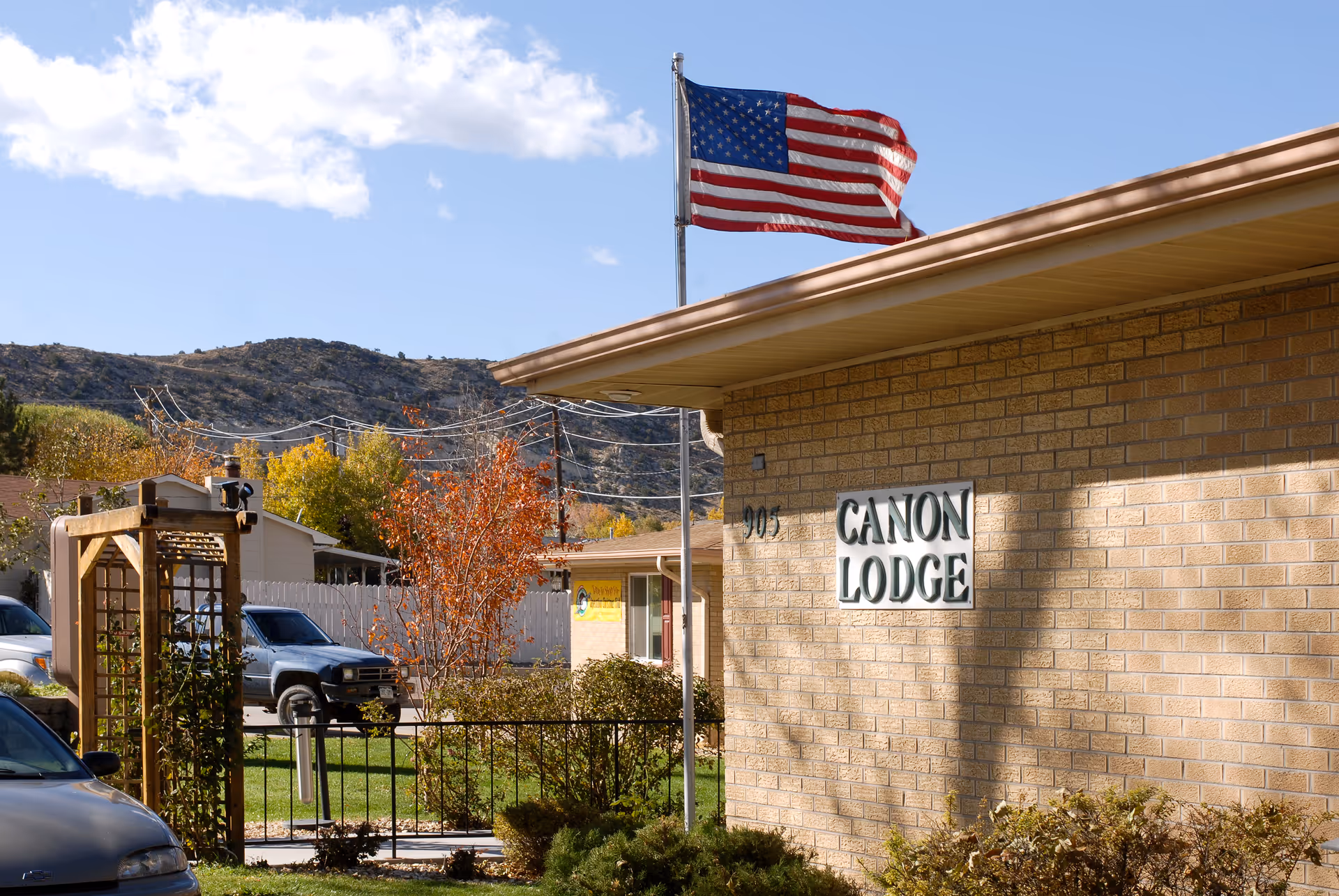 Exterior view of Canon Lodge Care Center building with a brick facade, an American flag on a flagpole, some bushes, a small tree with autumn leaves, parked cars, and hills in the background under a partly cloudy sky.