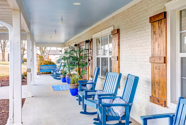 A covered outdoor porch area at Legacy Village of Hendersonville featuring blue rocking chairs with patterned cushions, potted plants, wooden window shutters, and a blue bench in the background. The porch overlooks a grassy area with trees.