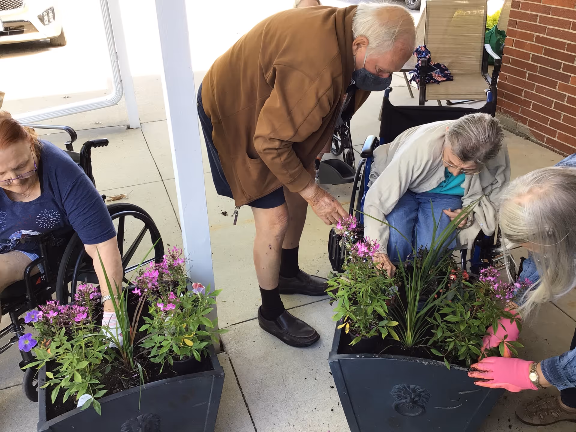 Four elderly individuals, three in wheelchairs and one standing, are engaged in planting and tending to flowers in two large black planters on a concrete patio outside a brick building. One person is wearing pink gardening gloves, and the standing person is wearing a face mask.