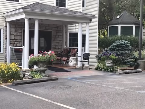 Entrance of a building with a covered porch supported by white columns. There are two cushioned chairs and a small table on the porch, along with large planters filled with flowers and greenery. To the right, there is a gazebo surrounded by trees and shrubs. The foreground shows a paved parking area with marked parking spaces.