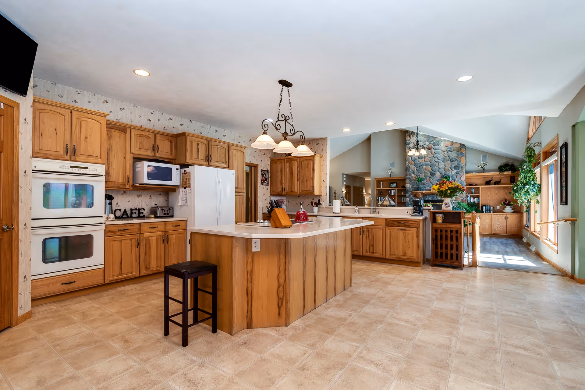 Spacious kitchen with wooden cabinets, a large island with a stool, white double oven, microwave, and refrigerator. The kitchen opens into a living area with a stone fireplace, built-in wooden shelves, and large windows letting in natural light.