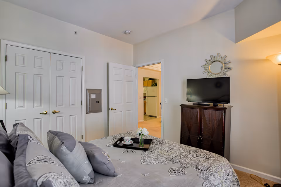 A bedroom with a bed covered in a patterned gray and white comforter and multiple pillows. A tray with a teacup and a small vase with white flowers sits on the bed. There are two closed white closet doors on the left wall and an open door leading to a kitchen area with a refrigerator. A dark wooden cabinet with a flat-screen TV on top is against the right wall, with a decorative mirror above it. A floor lamp is partially visible in the corner.