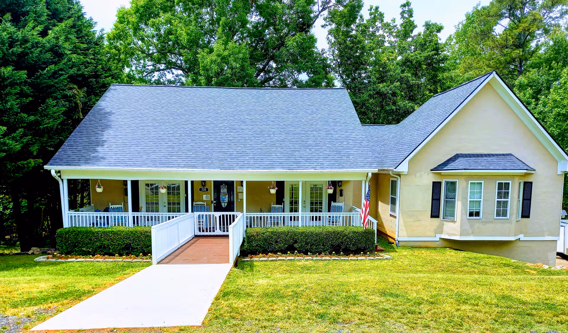 Front exterior view of a single-story building with a gray shingled roof, beige walls, and a covered porch with white railings and rocking chairs. There is a ramp leading up to the porch, green bushes and flowers along the front, and an American flag on the right side. Tall green trees are visible in the background.
