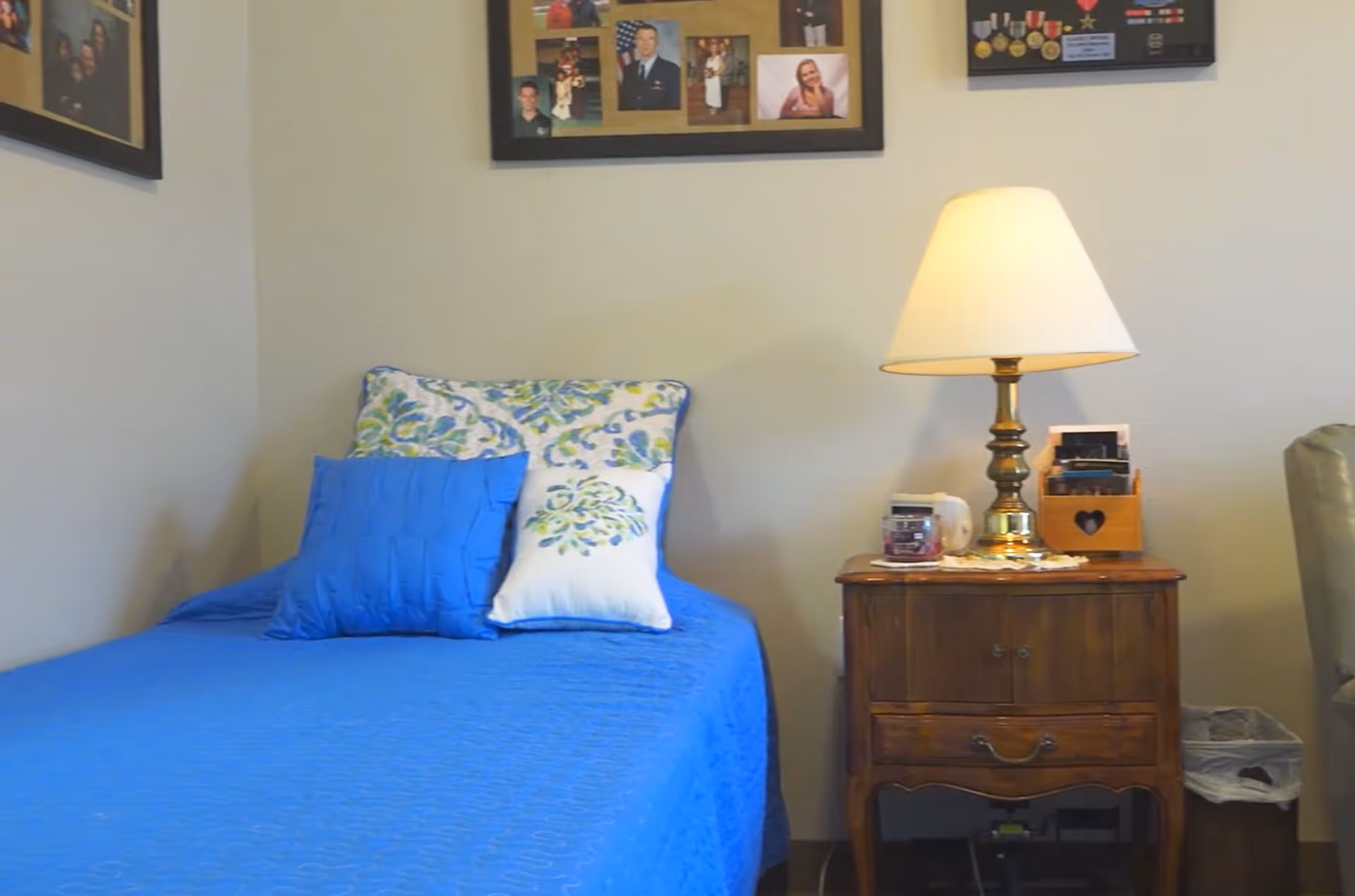 A bedroom with a single bed covered in a blue bedspread and two decorative pillows, one blue and one white with a floral pattern. Next to the bed is a wooden nightstand with a brass lamp, a candle, and a wooden organizer. The wall behind the bed has framed photos and memorabilia.