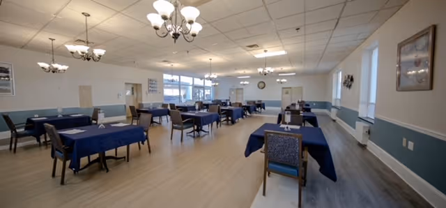 Spacious dining room with multiple tables covered in blue tablecloths, chairs, chandeliers, and large windows.