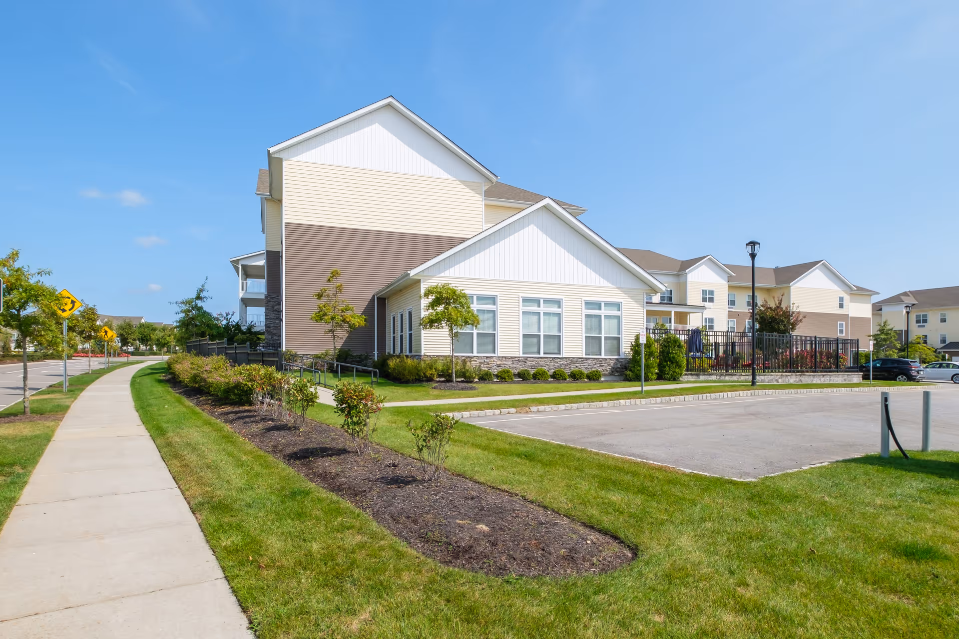 Exterior view of a senior living facility building with beige and brown siding, multiple windows, and a well-maintained lawn with small trees and shrubs. There is a sidewalk on the left side and a parking lot on the right under a clear blue sky.