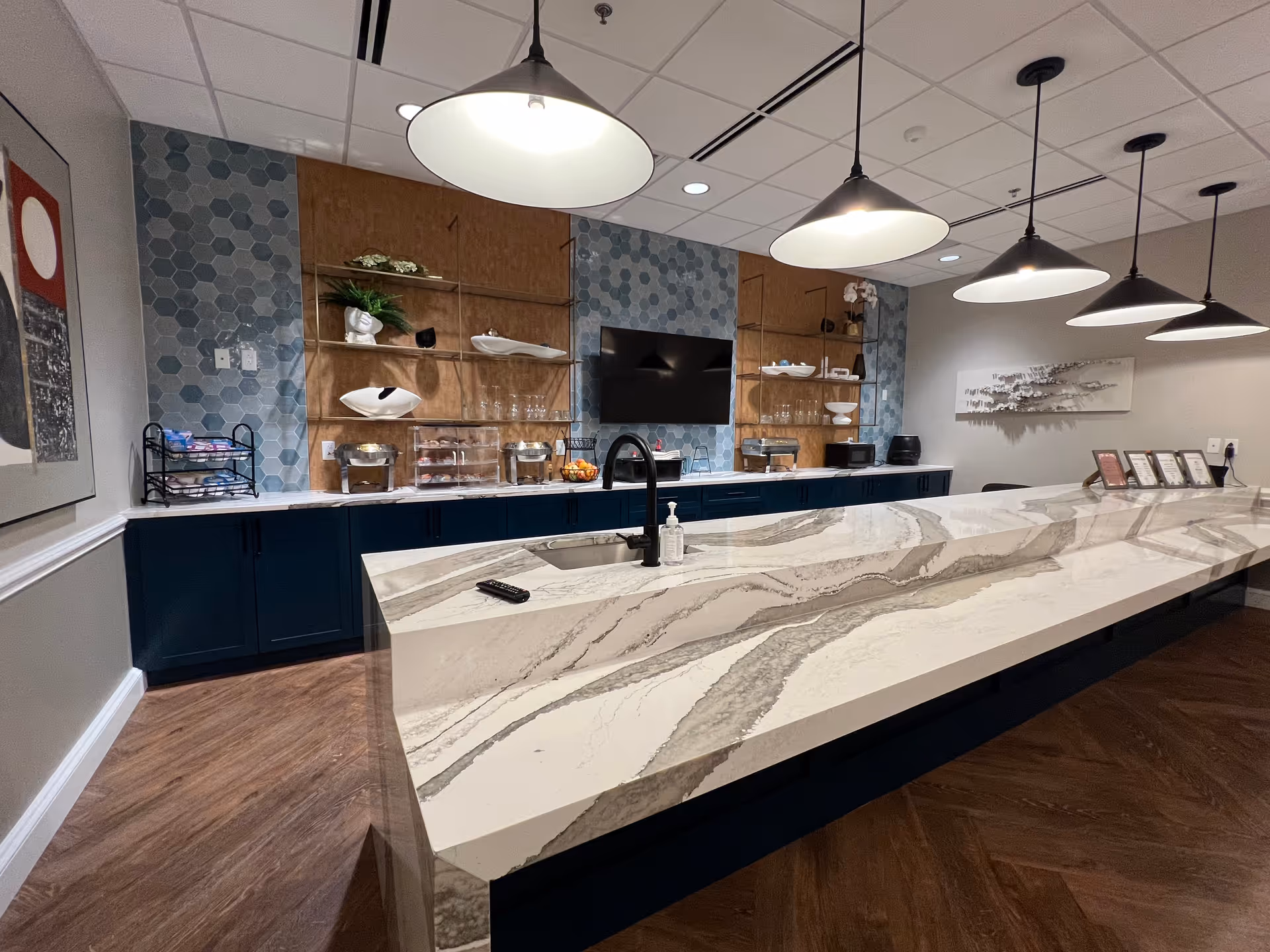 Modern kitchen area with a long marble countertop island featuring a sink and faucet, illuminated by five hanging pendant lights. The back wall has blue hexagonal tile accents, wooden shelves with decorative items, a flat-screen TV, and dark blue cabinets below. The floor is wood, and there is framed artwork on the left wall.