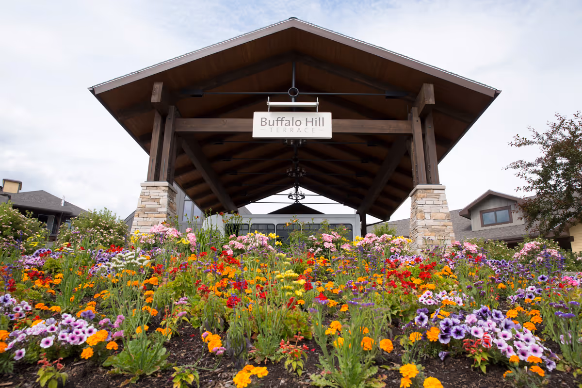 A wooden pavilion structure with stone pillars and a sign that reads 'Buffalo Hill Terrace' above the entrance. In front of the pavilion is a colorful flower garden with various blooming flowers including purple, orange, red, yellow, and pink blossoms. Residential buildings are visible in the background under a cloudy sky.