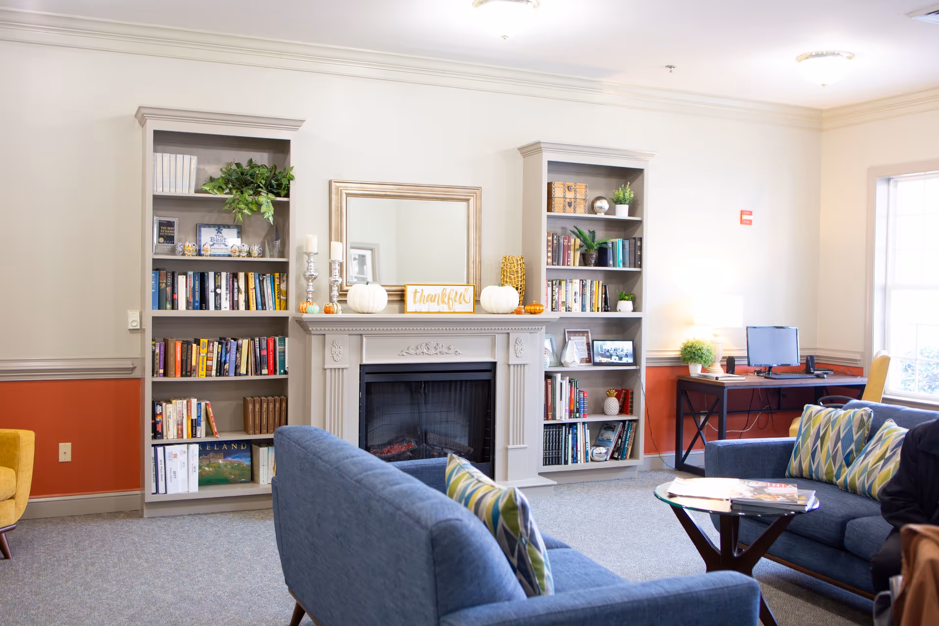 A cozy living room area with two blue sofas adorned with patterned pillows, a glass coffee table with magazines, a fireplace with decorative pumpkins and a 'thankful' sign, flanked by two tall bookshelves filled with books and plants. A desk with a computer and a lamp is positioned near a window letting in natural light.