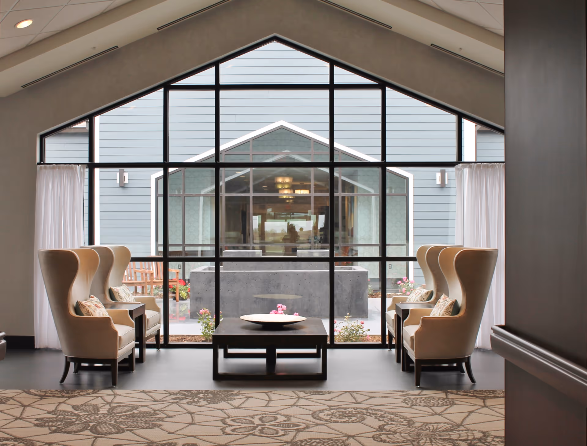 A cozy seating area with four beige wingback chairs arranged around a dark wooden coffee table, positioned in front of large floor-to-ceiling windows that look out onto an outdoor courtyard with flowers and a water feature.
