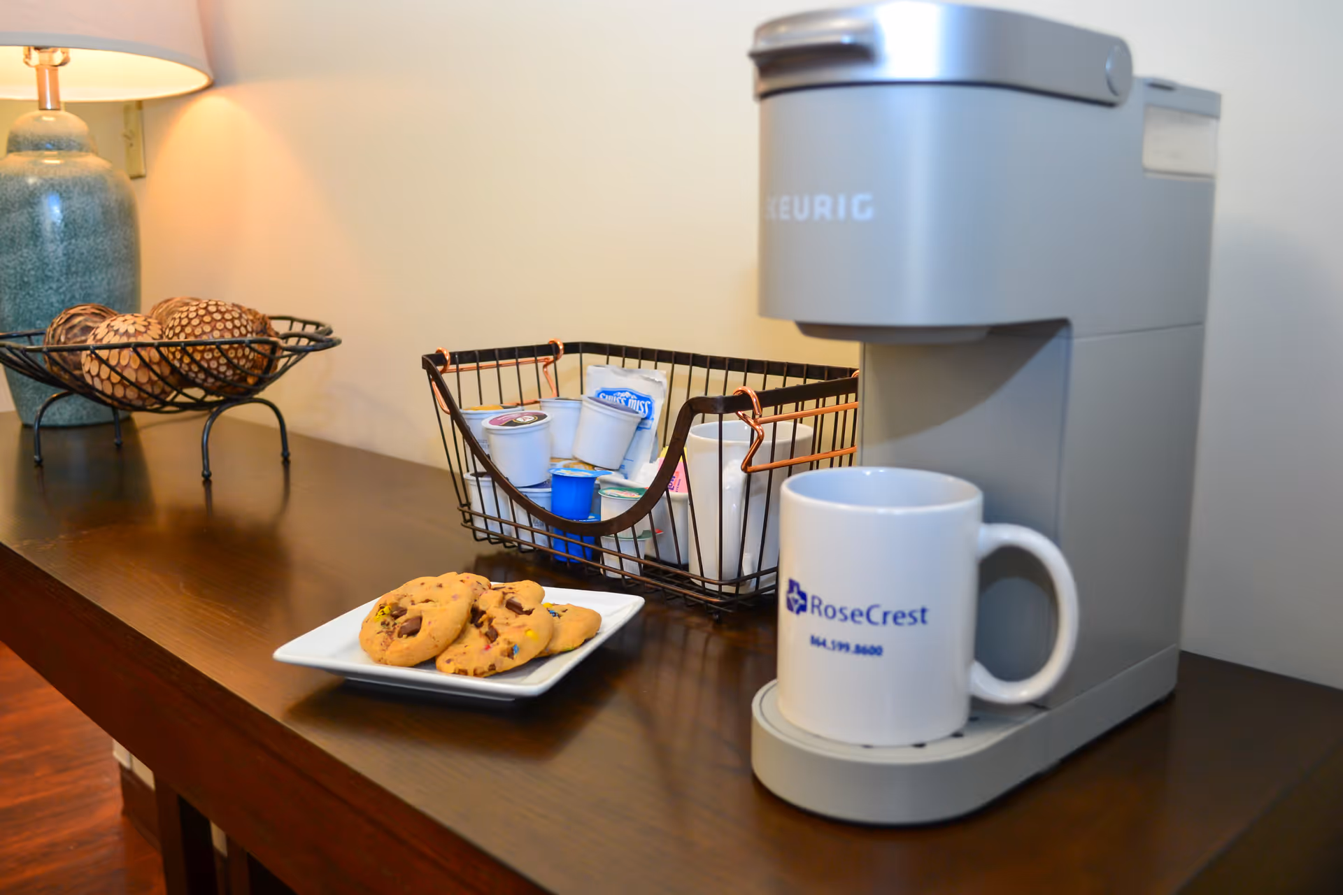 A coffee station on a wooden table featuring a Keurig coffee maker with a white RoseCrest mug, a plate with chocolate chip cookies, a wire basket holding coffee pods and creamers, and a green ceramic lamp with a beige lampshade.