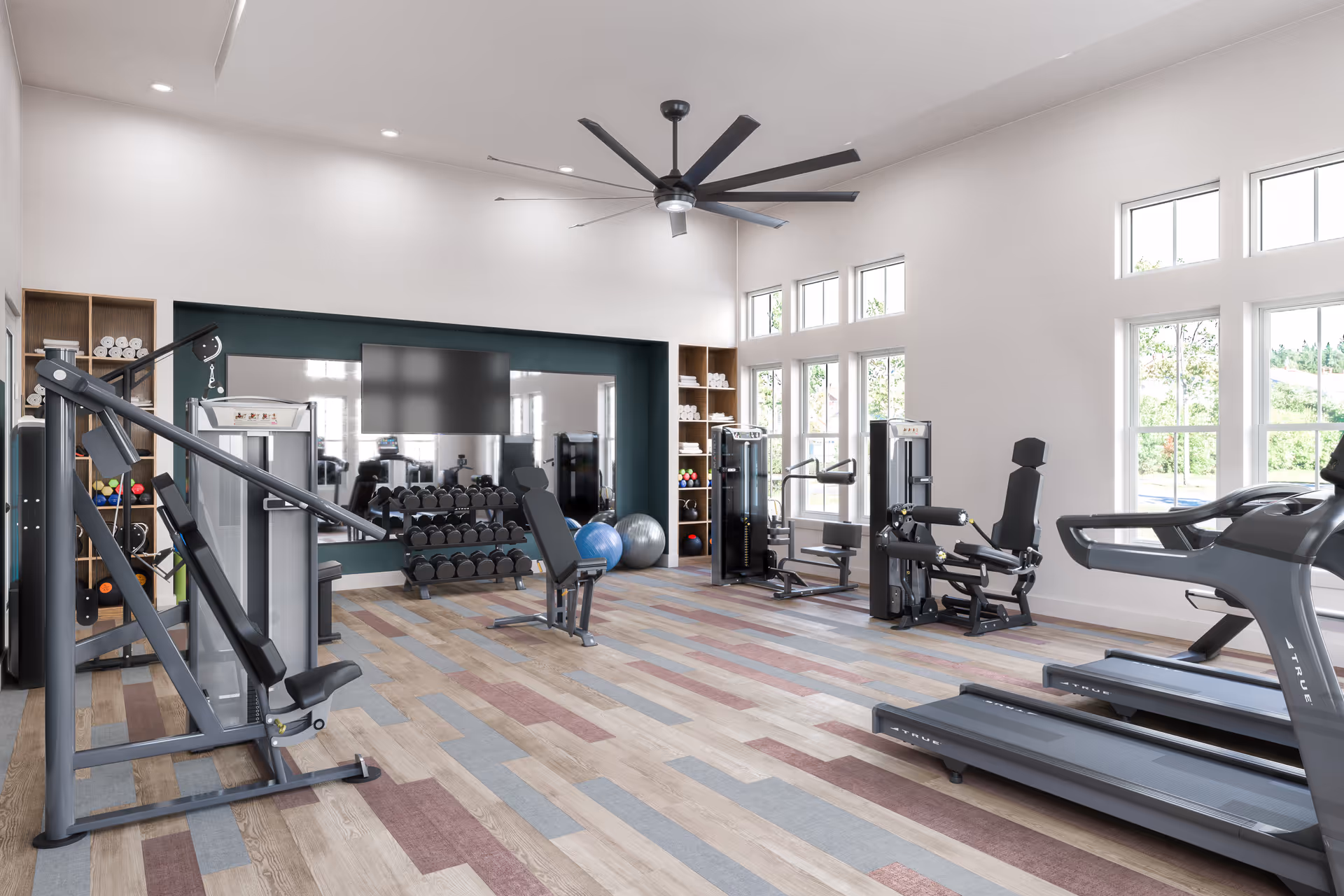 Bright and spacious fitness room with large windows letting in natural light. The room features various exercise equipment including treadmills, weight machines, dumbbells, exercise balls, and shelves stocked with towels and small weights. A large ceiling fan is mounted in the center of the ceiling, and a flat-screen TV is mounted on the wall above the dumbbell rack.