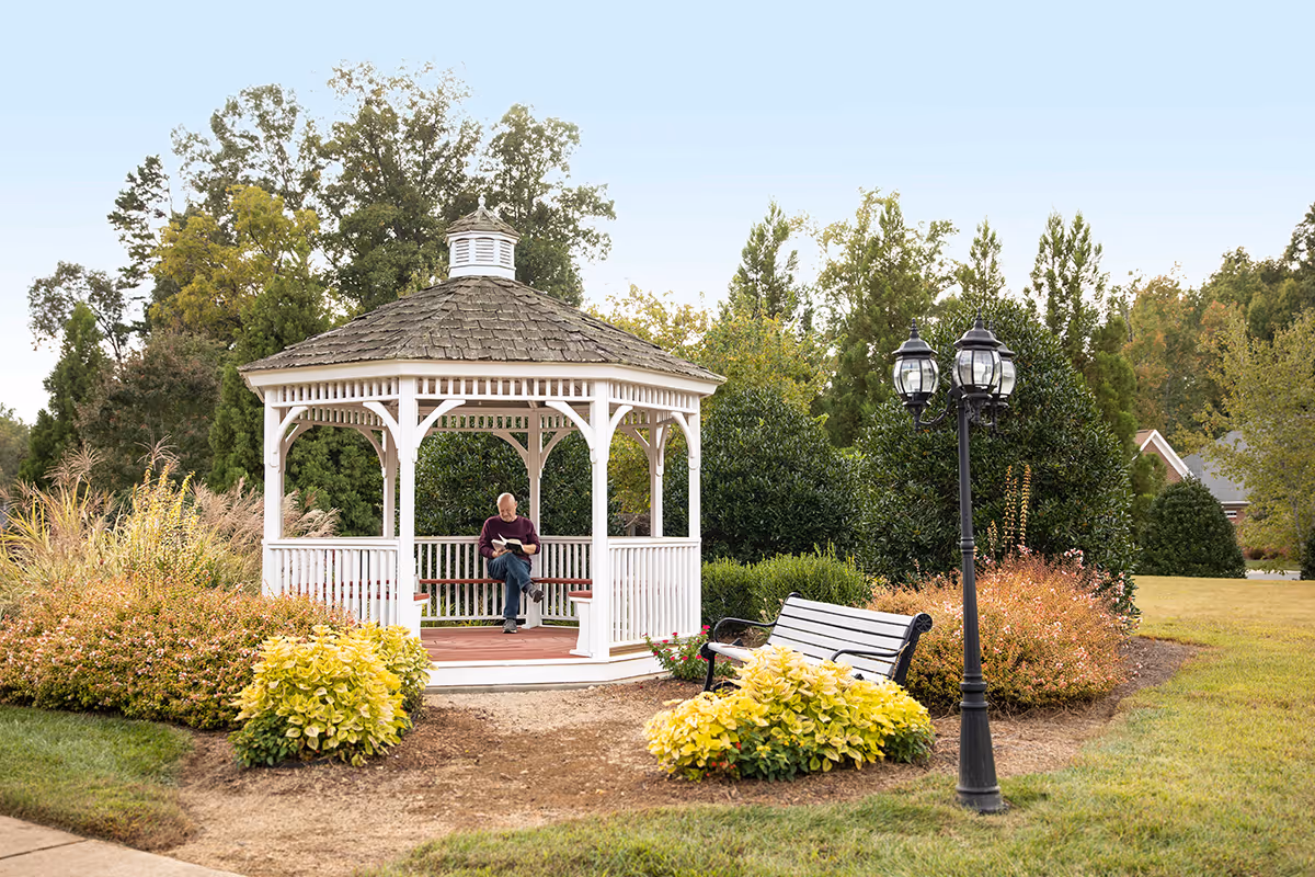 White wooden gazebo in a landscaped garden with a person sitting inside reading, a bench and a lamppost nearby.