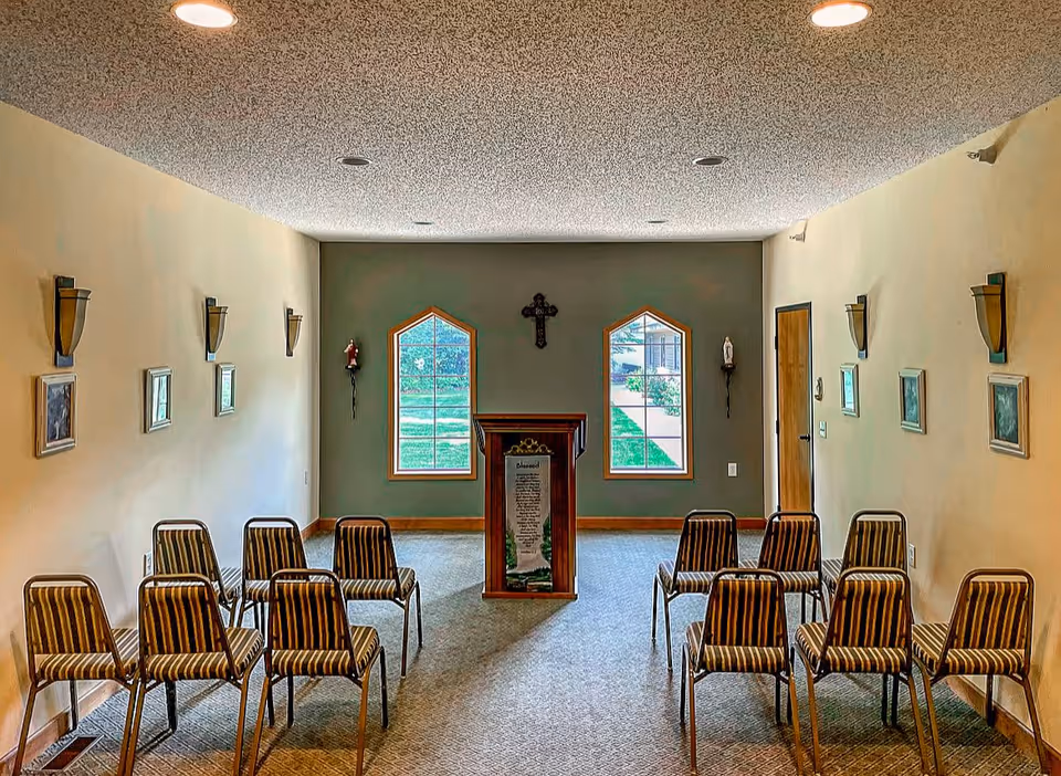 A small chapel or meditation room with two arched windows at the far end, a wooden podium with a religious text in the center, a cross mounted on the wall between the windows, and two statues on either side. There are striped chairs arranged in rows facing the podium, wall sconces, and framed pictures on the side walls.
