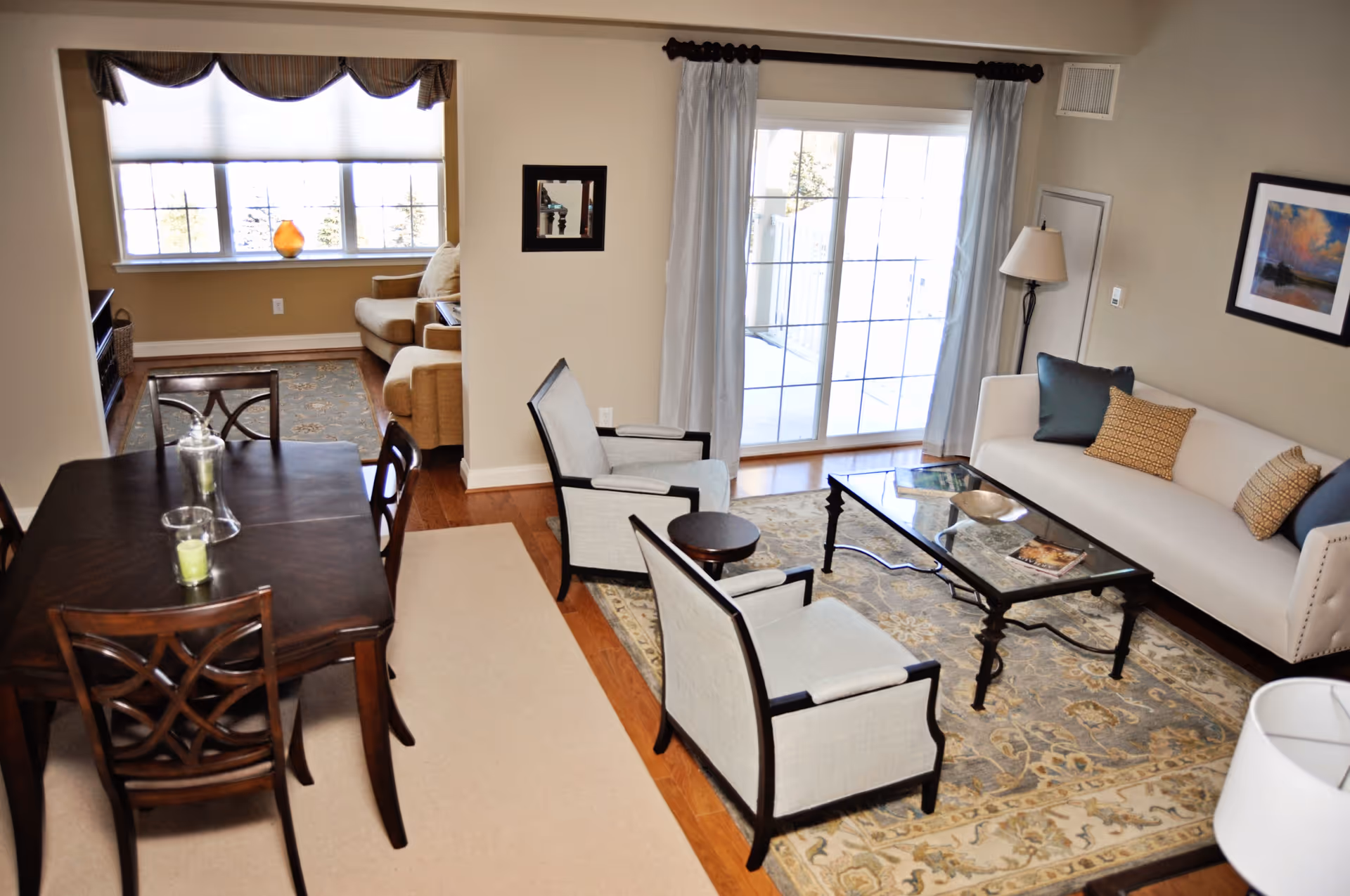 A bright living and dining area featuring a dark wooden dining table with chairs, a beige sofa with decorative pillows, two upholstered armchairs, a glass coffee table on a patterned rug, a floor lamp, and large windows with curtains allowing natural light to fill the room.