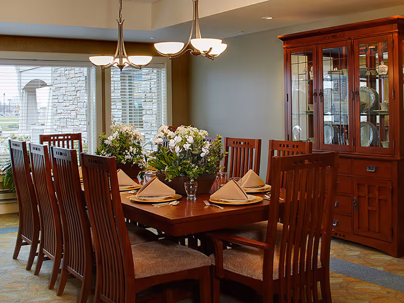 A formal dining room with a large wooden table set for eight people. The table is decorated with folded beige napkins, plates, glasses, and two floral centerpieces. The room has large windows with blinds, a wooden china cabinet displaying dishes, and two ceiling light fixtures.