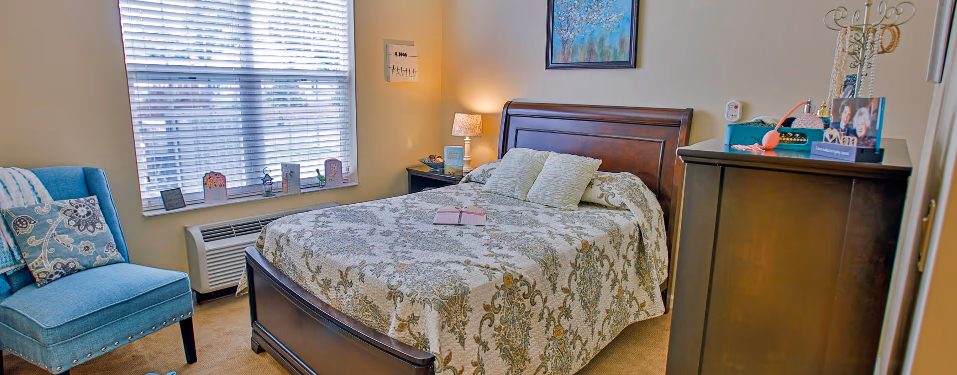 Sunlit bedroom with a patterned bed and wooden headboard, a dresser, and a blue upholstered chair by a window.
