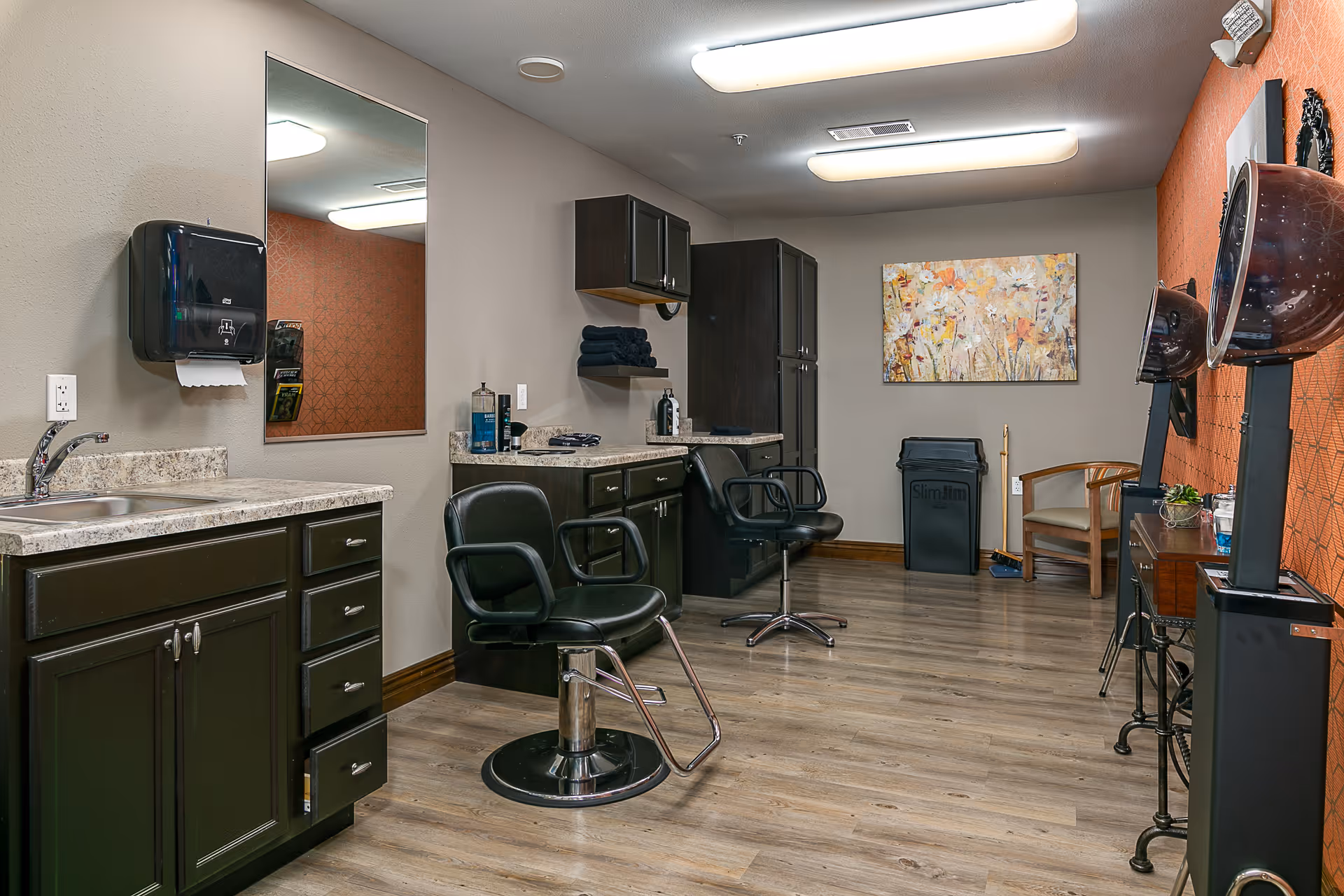 Interior view of a salon room with two black salon chairs, a large mirror, dark wood cabinets, a countertop with hair care products, and two hair dryers mounted on stands. The room has wood flooring, a beige wall with a colorful floral painting, and an orange accent wall with a geometric pattern.