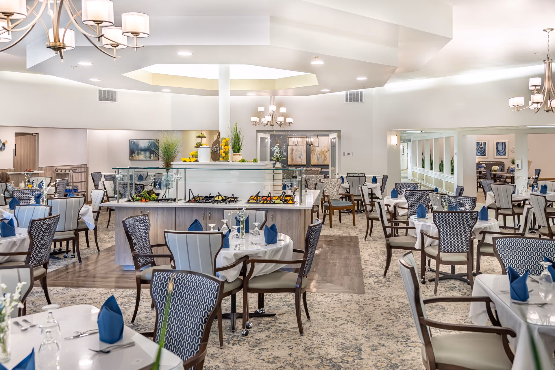 A spacious and well-lit dining room in a retirement residence with multiple round tables covered with white tablecloths, each set with blue napkins, glassware, and silverware. The room features comfortable chairs with patterned upholstery, modern chandeliers, and a central buffet area with food trays and decorative plants. The overall decor is elegant and inviting.