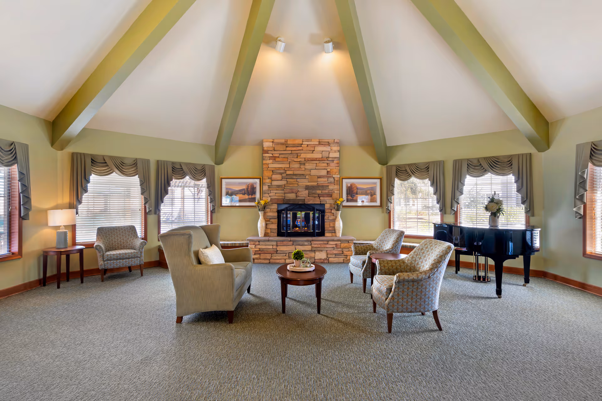 A spacious living room with a high vaulted ceiling featuring exposed beams. The room has a stone fireplace centered on the far wall, flanked by two framed pictures and vases with flowers. There are four upholstered armchairs arranged around a small round wooden coffee table in the middle of the room. Large windows with decorative valances allow natural light to fill the space. A black grand piano is positioned near the windows on the right side of the room.