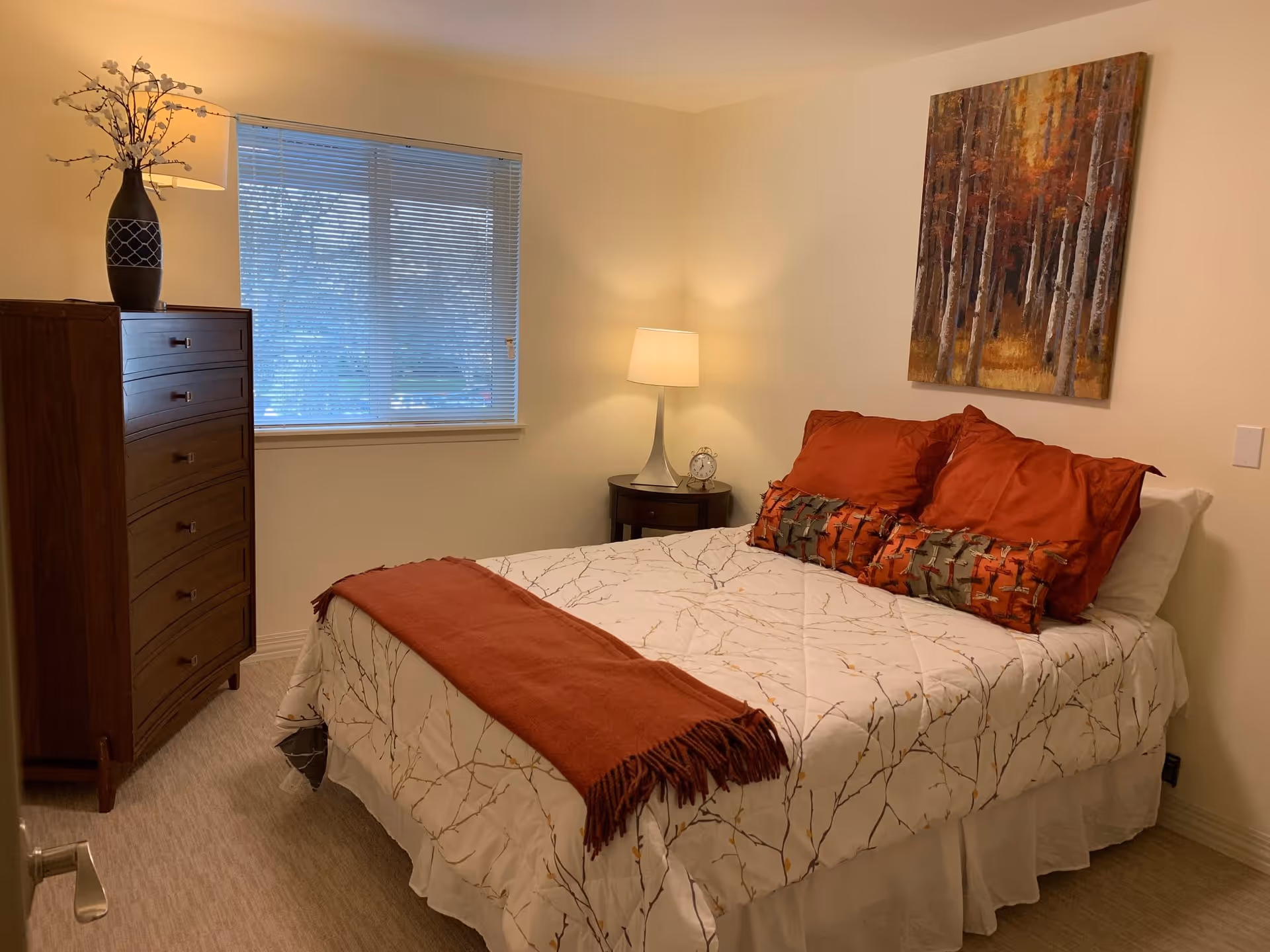 A cozy bedroom with a bed dressed in white bedding with a branch pattern, rust-colored pillows, and a matching throw blanket. There is a wooden dresser with a decorative vase and lamp on top, a window with blinds, a round bedside table with a lamp and an alarm clock, and a painting of a forest on the wall.