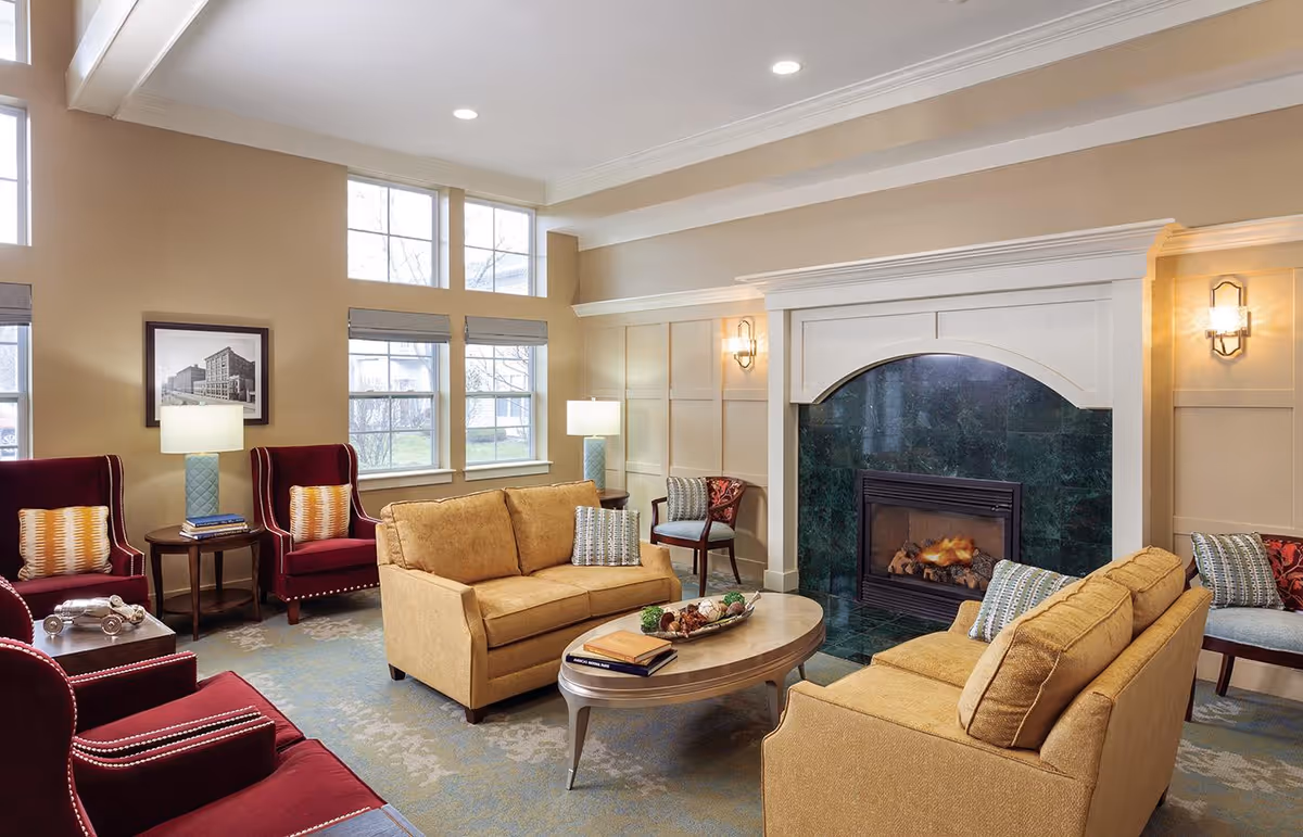 Bright communal living room with sofas, armchairs, a coffee table and a lit fireplace under large windows.