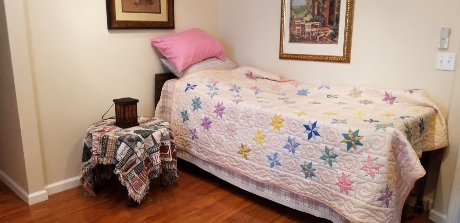 A small bedroom with a single bed covered by a quilt featuring colorful star patterns. There is a pink pillow on the bed. Next to the bed is a small table draped with a patterned cloth and a decorative lantern on top. The walls are light-colored and decorated with framed artwork. The floor is wooden.