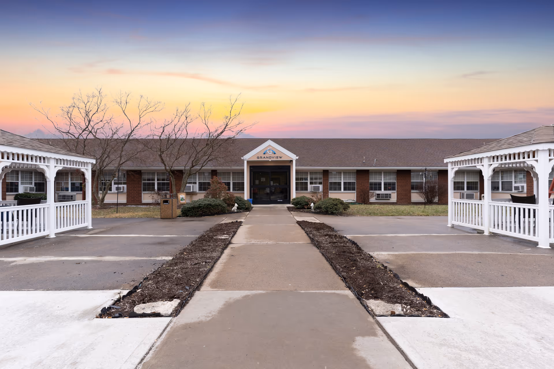 Front entrance of the Grandview Nursing & Rehabilitation building with a central walkway flanked by white gazebo structures and a colorful sunset sky.