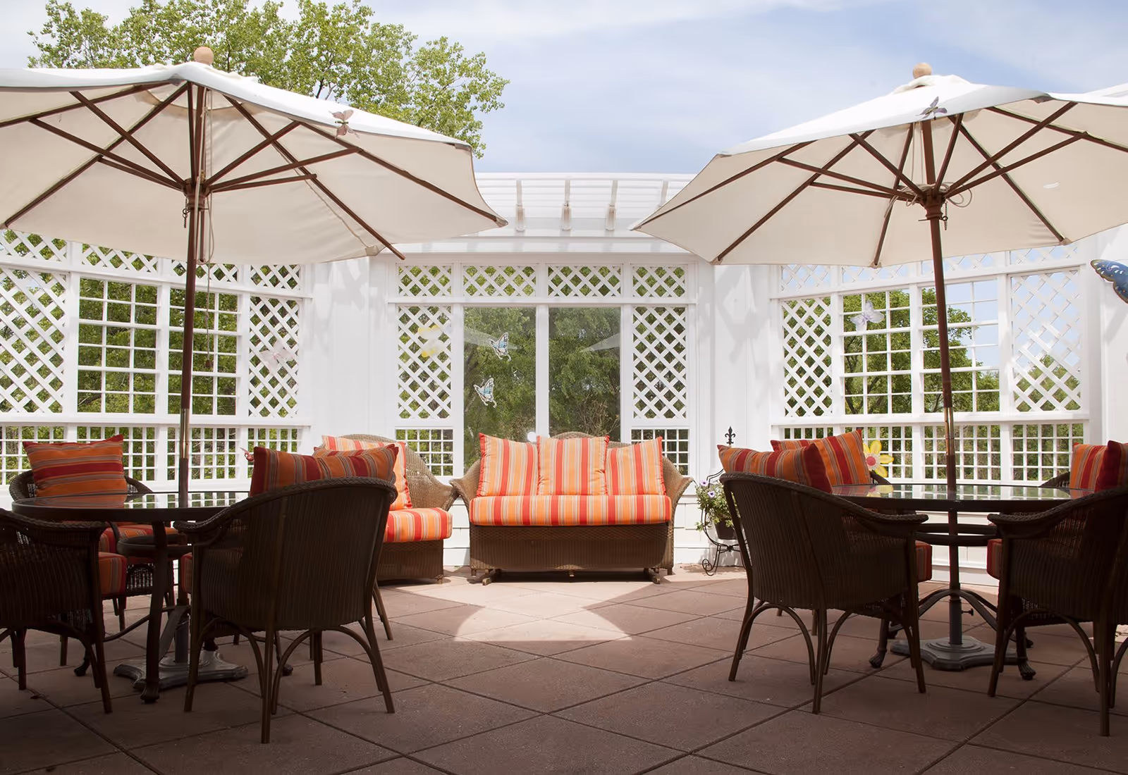 Outdoor patio area with two large white umbrellas shading tables and wicker chairs with red and orange striped cushions. A wicker loveseat with matching cushions is centered against a white lattice fence with glass doors and green trees visible outside.