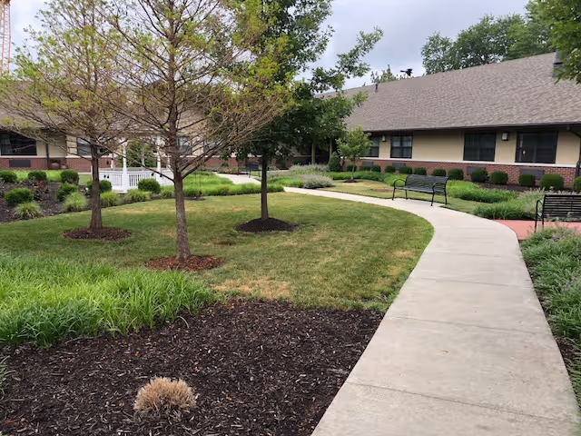 Paved walkway through a landscaped courtyard with benches, trees, and a single-story senior living building in the background.