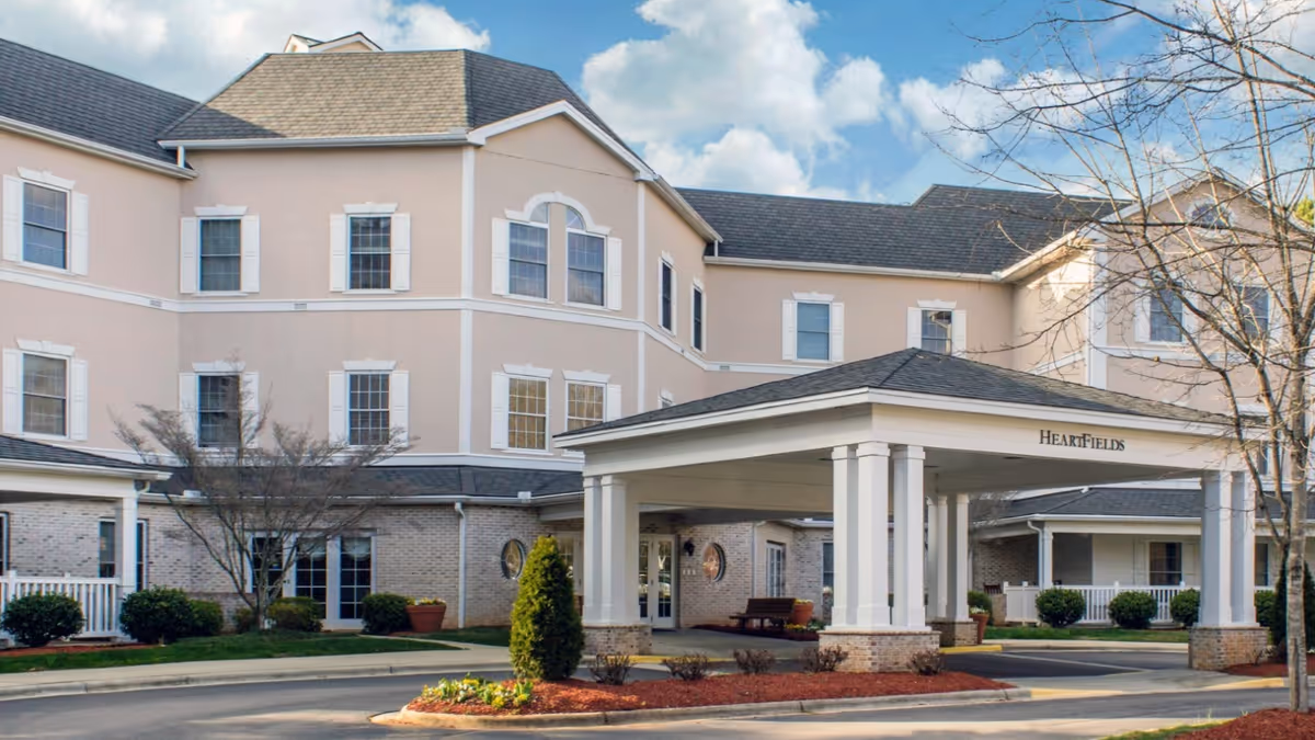 Exterior view of HeartFields at Cary senior living facility showing a large covered entrance with white columns, beige and brick building facade, multiple windows, landscaped bushes, and a tree without leaves under a partly cloudy sky.