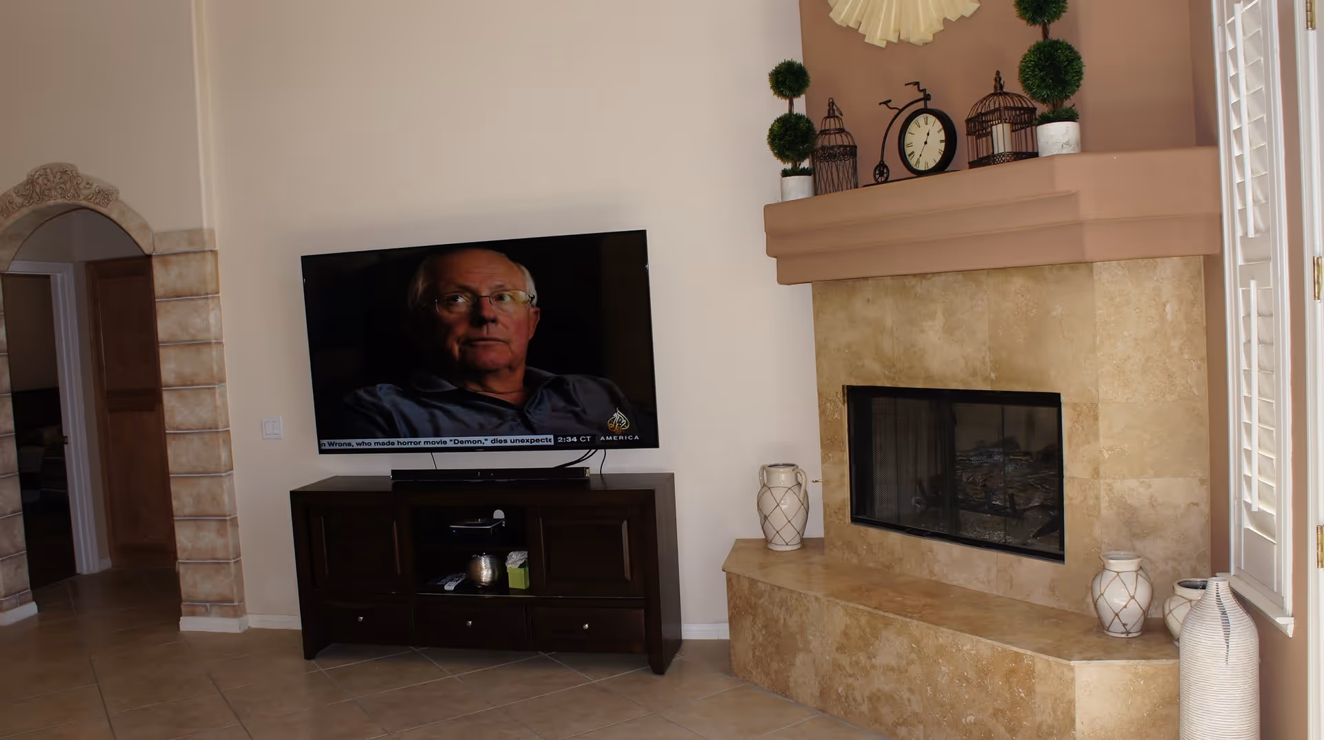A living room with a mounted flat-screen TV displaying a man, a dark wooden TV stand with decorative items, a beige tiled fireplace with various decorative plants and vases on the mantel and hearth, and an arched doorway with stone trim leading to another room.