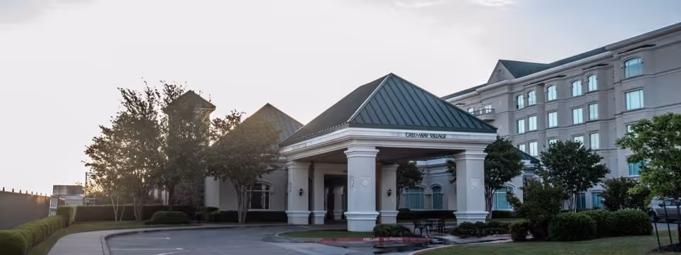 Exterior view of a senior living facility with a covered entrance supported by white columns, a green metal roof, and surrounding trees and bushes. The building has multiple stories with large windows and a parking area visible to the right.
