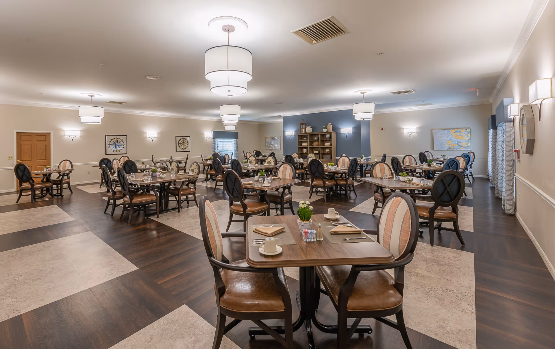 A spacious dining room with multiple wooden tables and cushioned chairs arranged neatly. Each table is set with cups, napkins, and small decorative plants. The room features a mix of dark wood and light tile flooring, soft lighting from ceiling fixtures, and wall sconces. The walls are decorated with framed artwork and a shelving unit with decorative items.