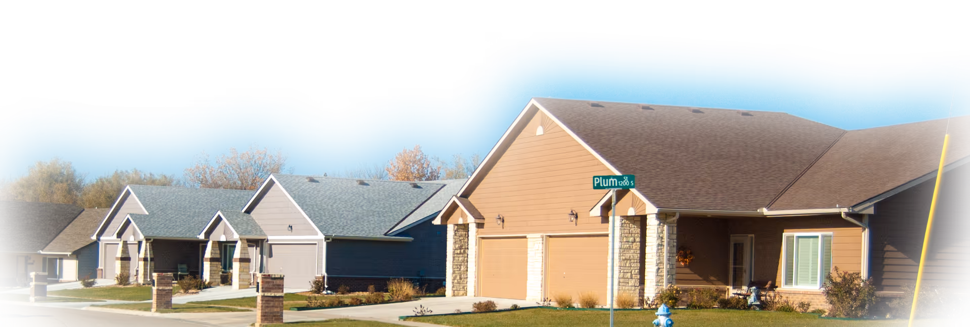 A suburban street view showing a row of single-story houses with garages, lawns, and driveways under a clear blue sky. A street sign labeled 'Plum' is visible near the corner house.