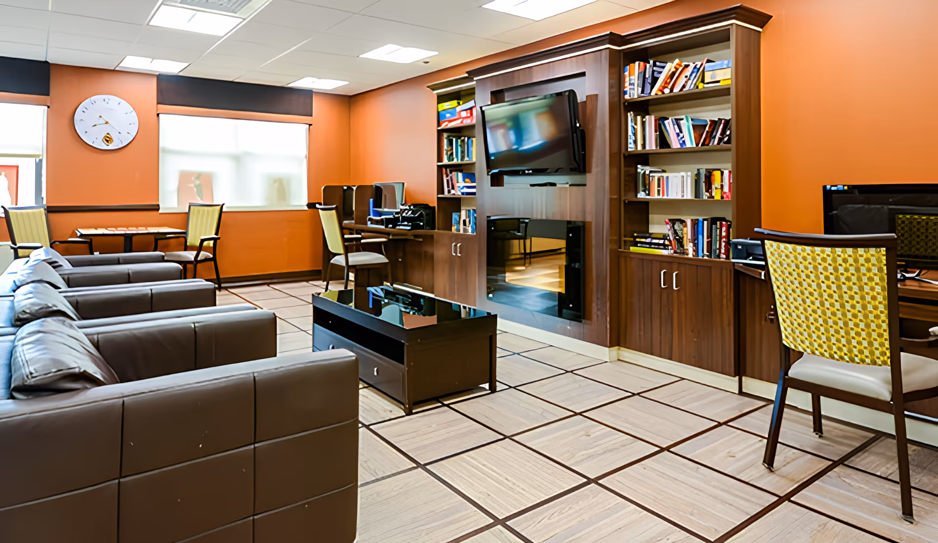 A cozy common area in a senior living facility with brown leather sofas, a wooden coffee table, a wall-mounted TV surrounded by bookshelves, and several chairs and desks along the walls. The walls are painted orange, and there is a large clock on one wall above two windows with blinds.