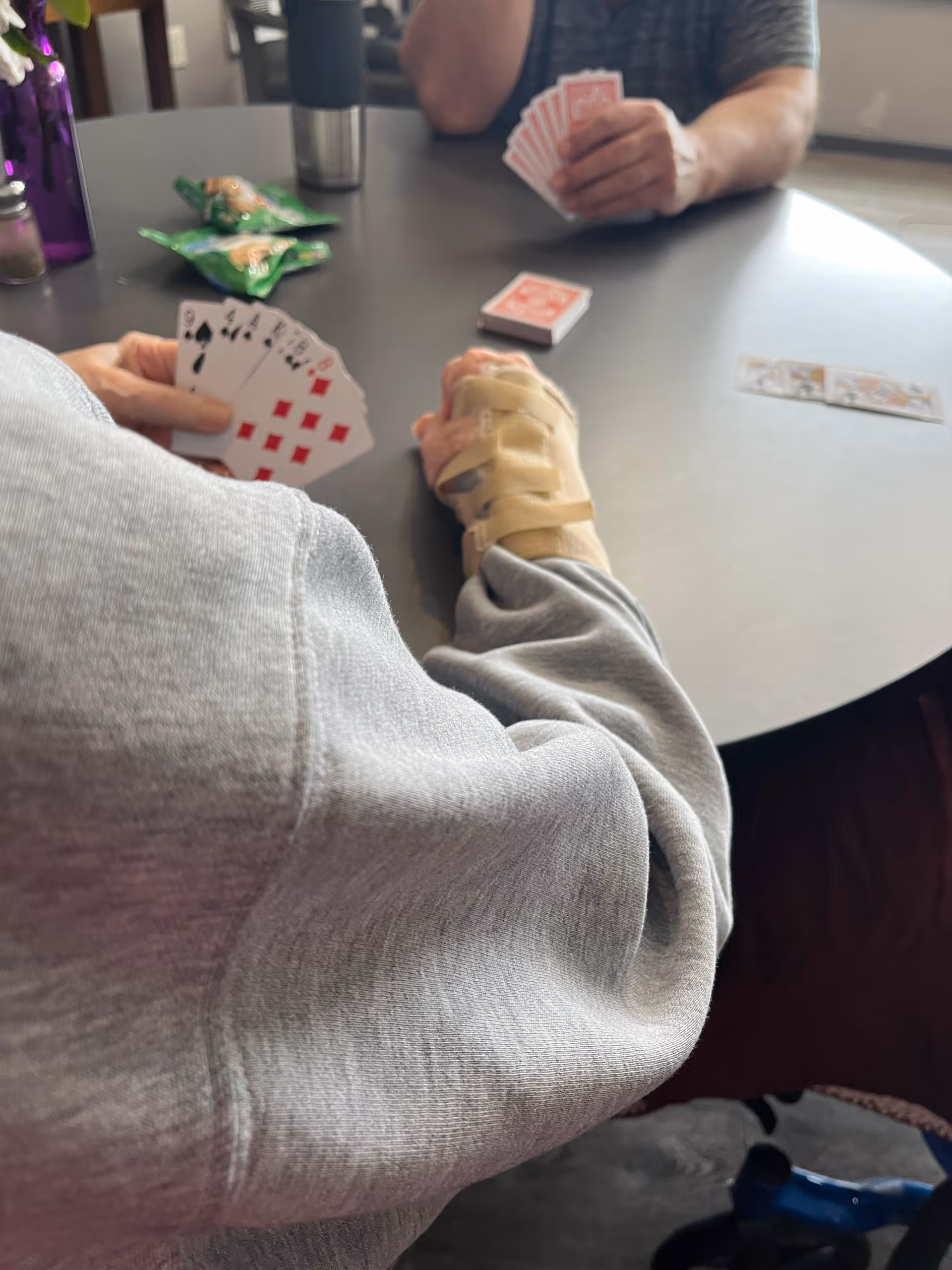 Two people sitting at a round table playing cards. One person is wearing a gray sweatshirt and has a wrist brace on their left hand. The other person is wearing a dark shirt. There are snacks and a stainless steel tumbler on the table.