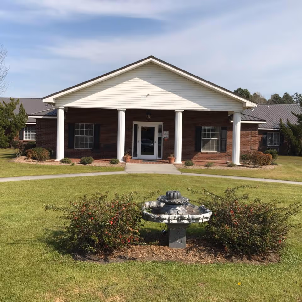 Front entrance of a single-story brick building with white columns and a covered portico, with a small fountain and lawn in the foreground.