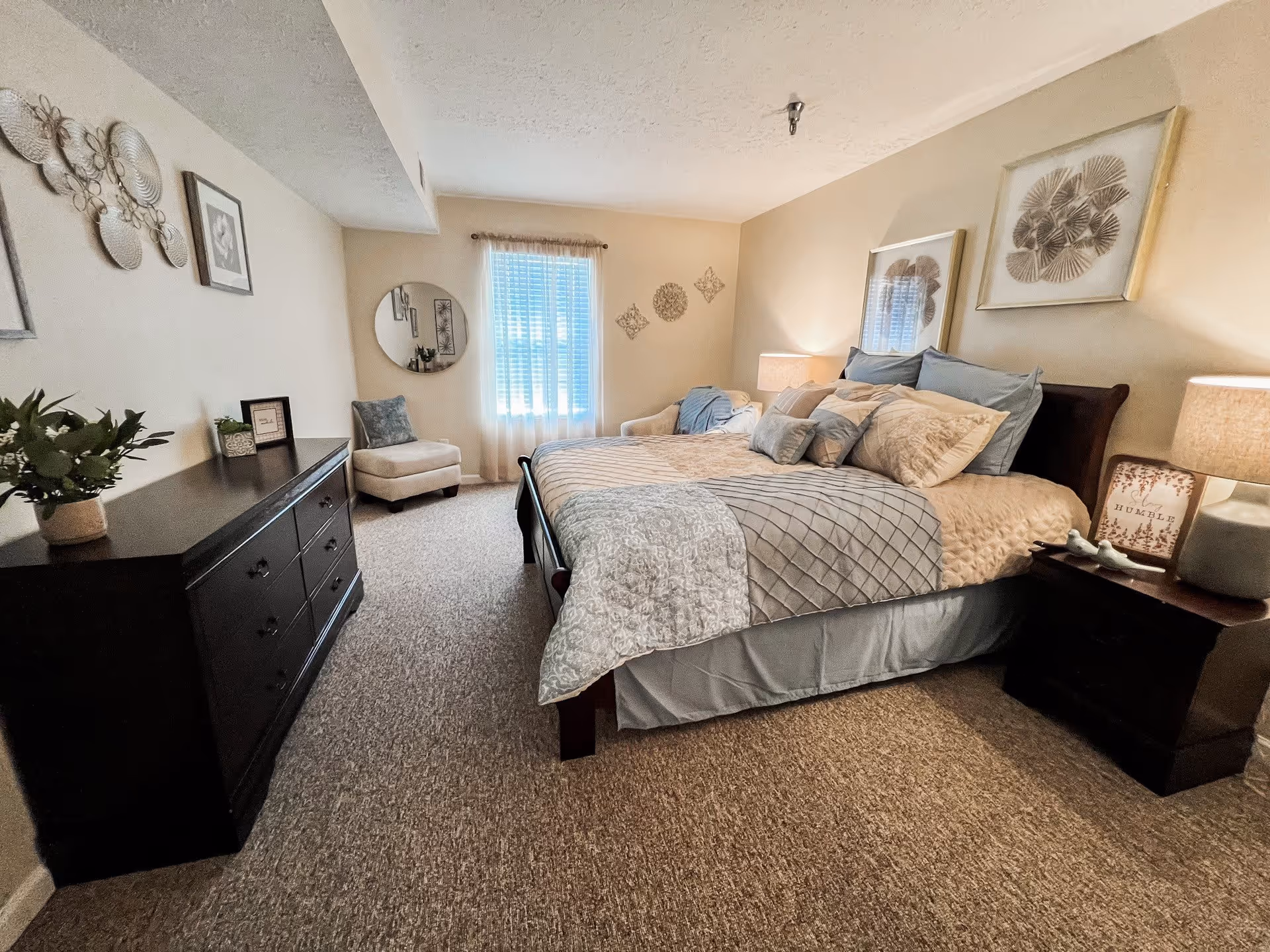 A cozy bedroom with a large bed covered in light blue and beige bedding, flanked by two dark wooden nightstands with lamps. There is a dark wooden dresser on the left side with a potted plant and framed pictures. A beige armchair is positioned near a window with sheer curtains. The walls are decorated with framed artwork and metal wall decor.