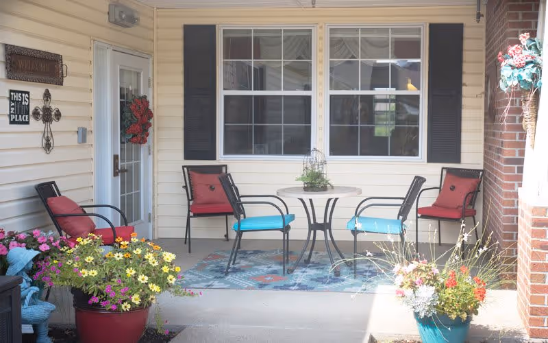 Covered front porch with a small round table, four chairs with colorful cushions, and potted flowers by a windowed entrance.