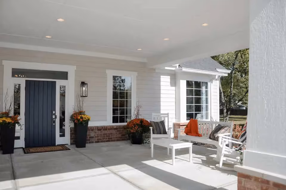 Covered front porch area of a house with a dark blue front door, two tall black planters with orange and yellow flowers on either side of the door, a white bench with cushions and an orange blanket, a white chair, and a small white table. The porch has a concrete floor and white siding with brick accents. Trees and grass are visible in the background.