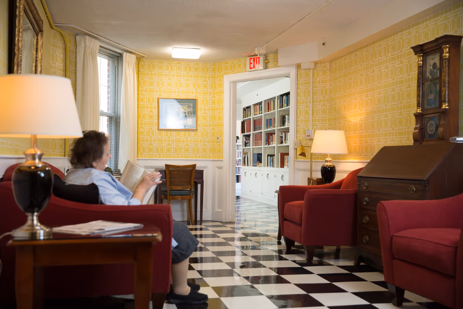 A cozy sitting area with red armchairs and a woman reading a book on one of the chairs. The room has yellow patterned wallpaper, black and white checkered floor tiles, two table lamps, a wooden side table, a wooden desk with drawers, and a grandfather clock. Through an open doorway, a room with white bookshelves filled with books is visible.