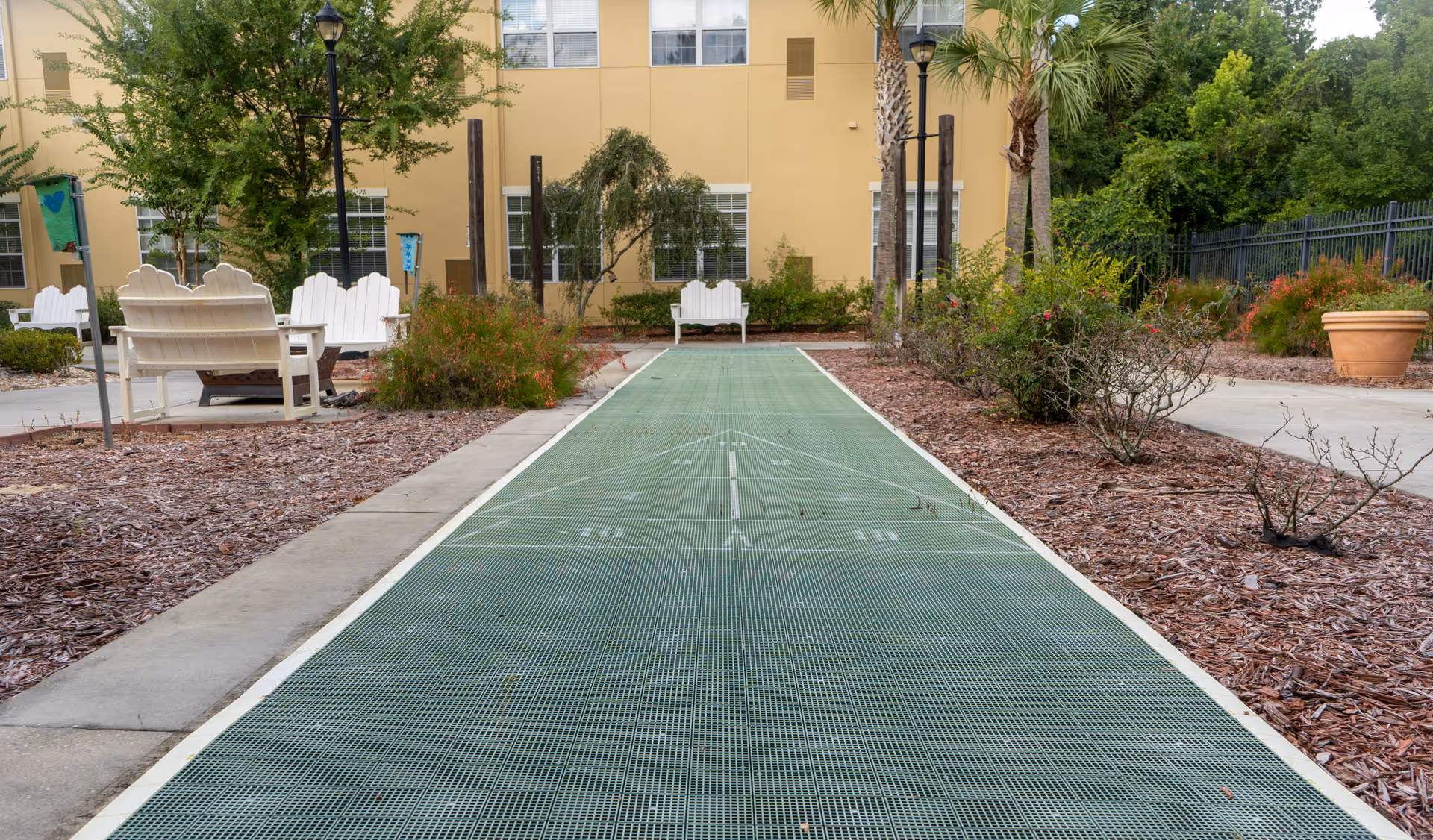 Outdoor shuffleboard court with green playing surface surrounded by mulch beds and bushes. White wooden benches and chairs are placed around the area, with a yellow building and trees in the background.