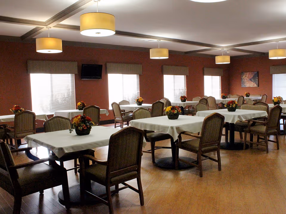 A dining room with multiple tables covered in white tablecloths, each adorned with a small floral centerpiece. The room has wooden floors, several windows with blinds and valances, and ceiling lights with round beige lampshades. Chairs with cushioned seats and backs surround the tables.