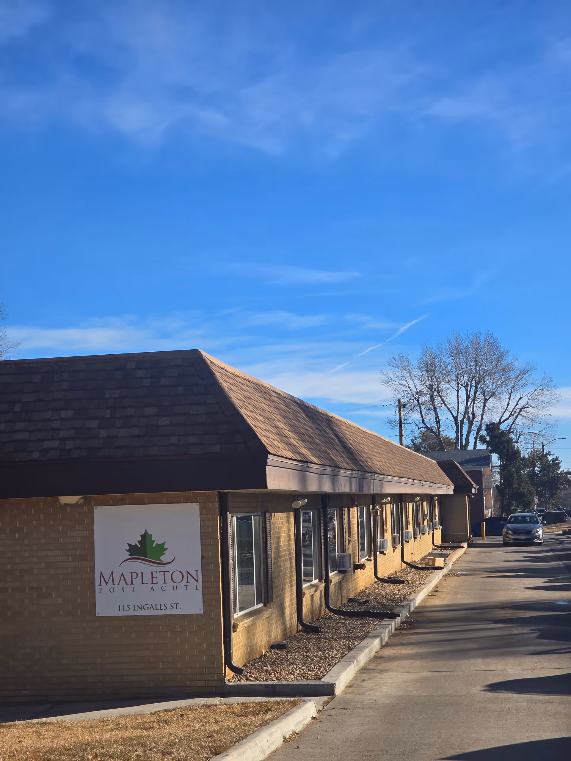 Exterior view of Mapleton Post Acute building with a brown shingled roof and brick walls under a clear blue sky. A sign on the building reads 'Mapleton Post Acute 115 Ingalls St.' There is a driveway alongside the building with a parked car in the distance and some leafless trees visible.