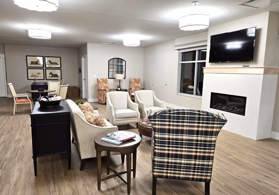 A cozy living room area in a senior living facility with a mix of upholstered chairs including a plaid armchair, a sofa, and two beige armchairs arranged around a small round table and a fireplace with a mounted flat-screen TV above it. The room has wood flooring, soft lighting from ceiling fixtures, and a window with a blind. In the background, there is a dining table with chairs and framed artwork on the wall.