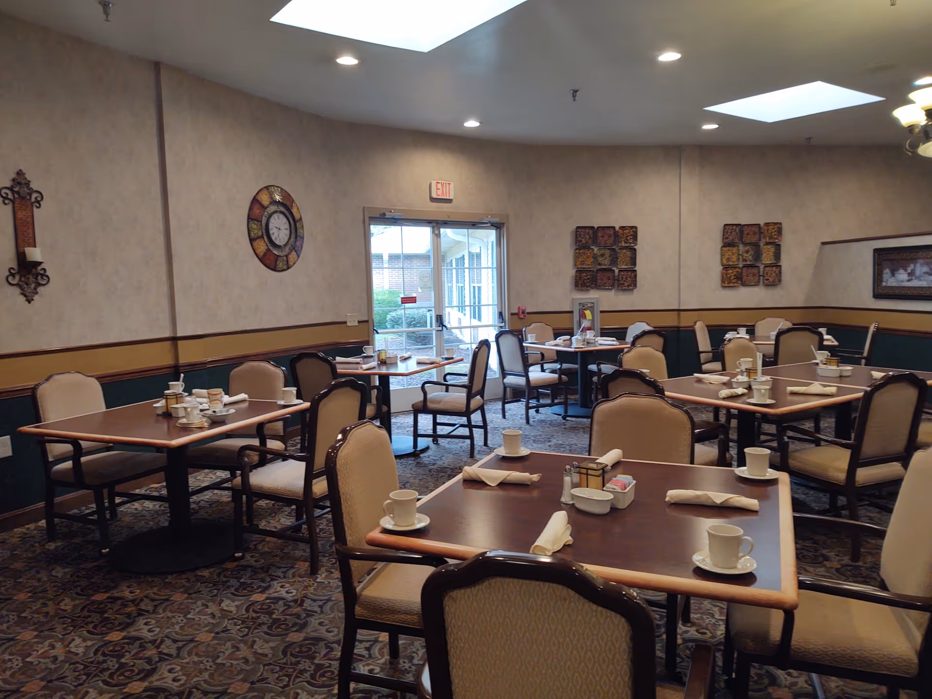 Dining room with multiple tables and chairs arranged neatly. Each table is set with white cups, saucers, napkins, and condiments. The room has patterned carpet, beige walls with decorative wall hangings, a clock, and a glass door leading outside. Ceiling lights and skylights provide illumination.