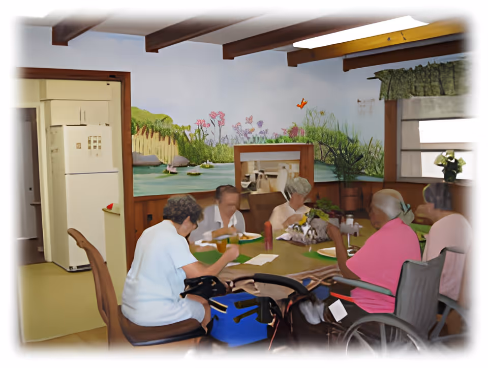 A group of elderly people sitting around a dining table in a room with wooden paneling and a mural of a garden scene on the wall. There is a refrigerator visible in an adjacent kitchen area, and one person is seated in a wheelchair.