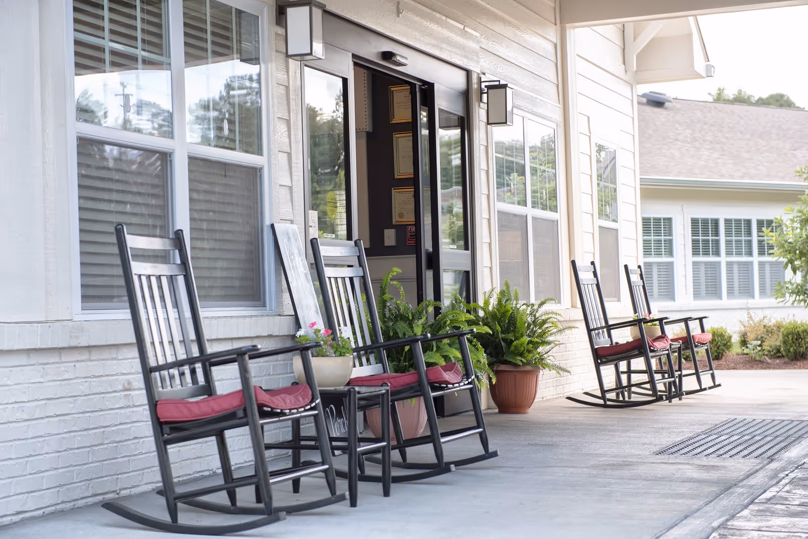 Front porch area of a senior living facility with several black rocking chairs with red cushions lined up along the wall. There are potted plants placed near the entrance door and windows with white blinds. The exterior walls are light-colored with white trim, and the entrance door is glass with a black frame.